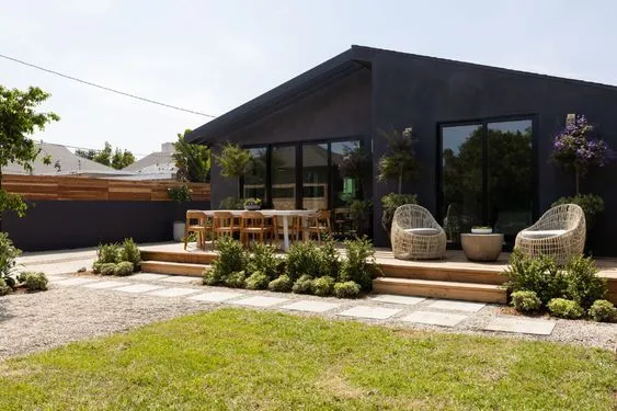 A black bungalow house design with lots of shrubs and plants and white patio furniture. 